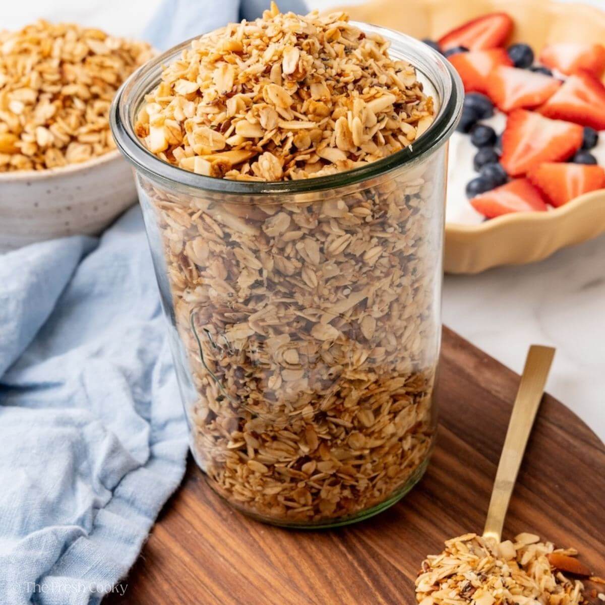 Soft-baked crock pot granola in a tall glass jar on a wooden board, with a bowl of granola, berries and a gold spoon nearby.