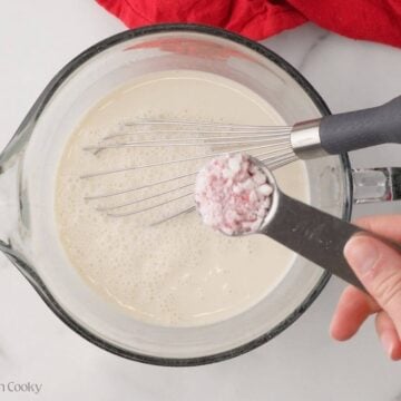 Hand holding crushed peppermint in a measuring spoon, hovering over the milk mixture.