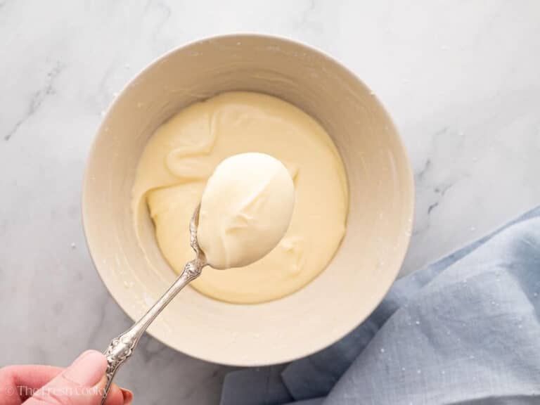 Cream cheese frosting in a white bowl. A hand lifting a spoonful of frosting upwards.