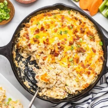 A cast iron skillet with spoon scooping out chicken bacon ranch dip and surrounded by veggies, scoops and dippers.