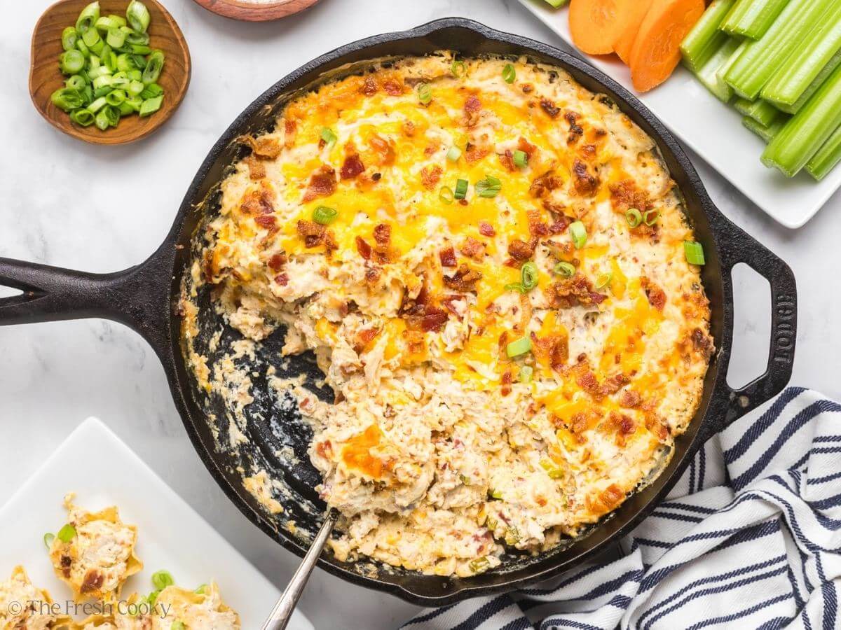A cast iron skillet with spoon scooping out chicken bacon ranch dip and surrounded by veggies, scoops and dippers.