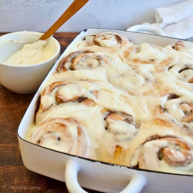 Gooey frosted cinnamon rolls in a pretty white enameled baking dish with frosting in a bowl.