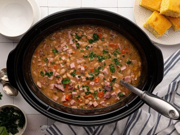 A black slow cooker bowl with ham and bean soup with a ladle in it and cornbread on the side.