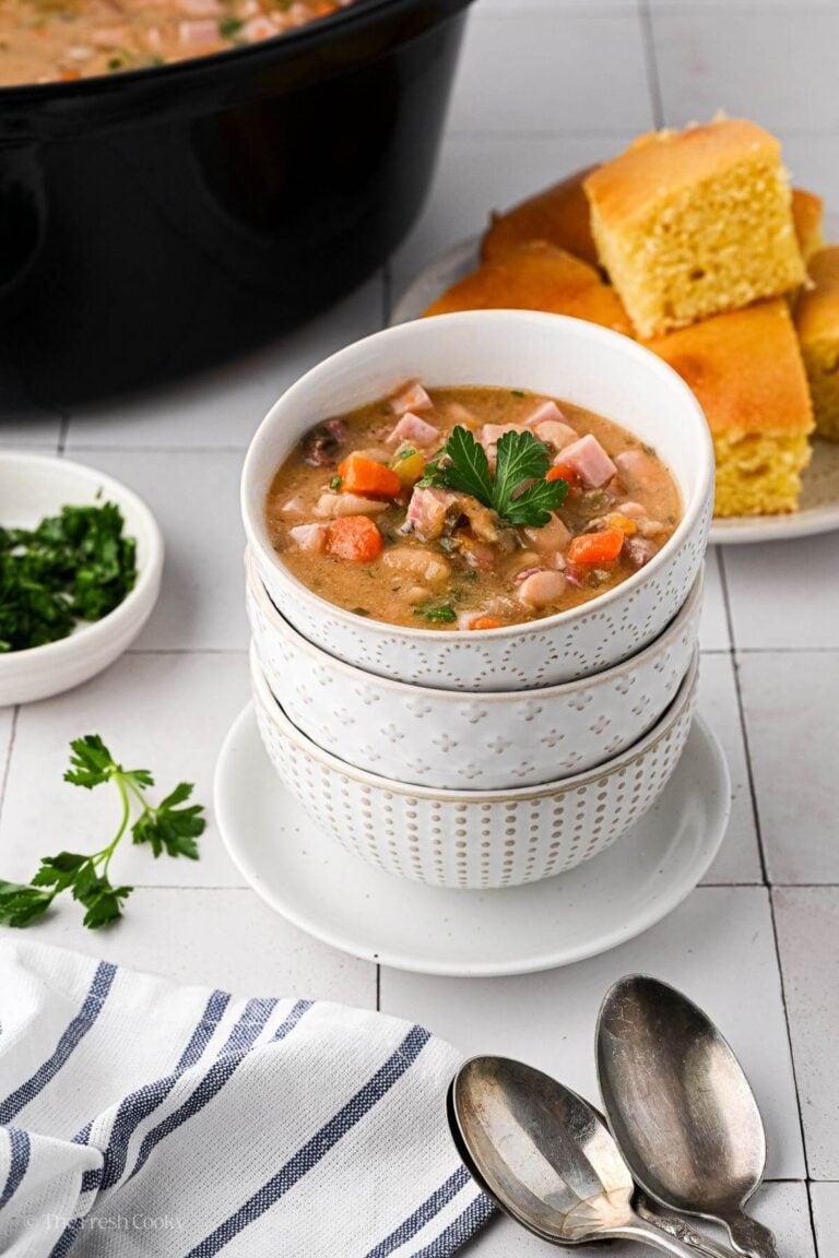 A stack of white bowls with slow cooker ham and bean soup. Cornbread is on a plate in the background.