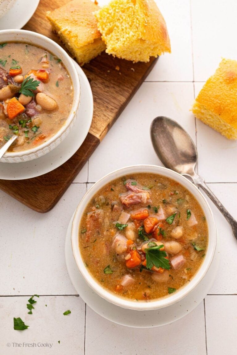 Two white bowls with slow cooker ham and bean soup with a spoon and cornbread.