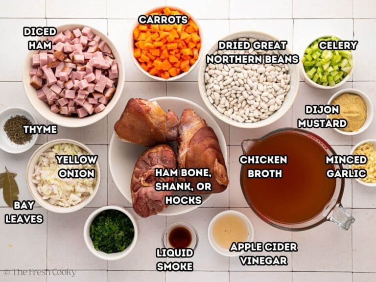 Ingredients for slow cooker ham and bean soup. L-R: Diced ham, thyme, bay leaf, yellow onions, carrots, ham bone, parsley, great northern beans, liquid smoke, chicken broth, apple cider vinegar, celery, mustard, and garlic.