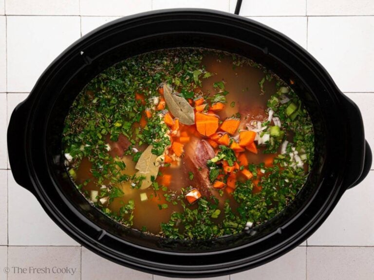 A black slow cooker bowl is filled with ingredients for slow cooker ham and bean soup.
