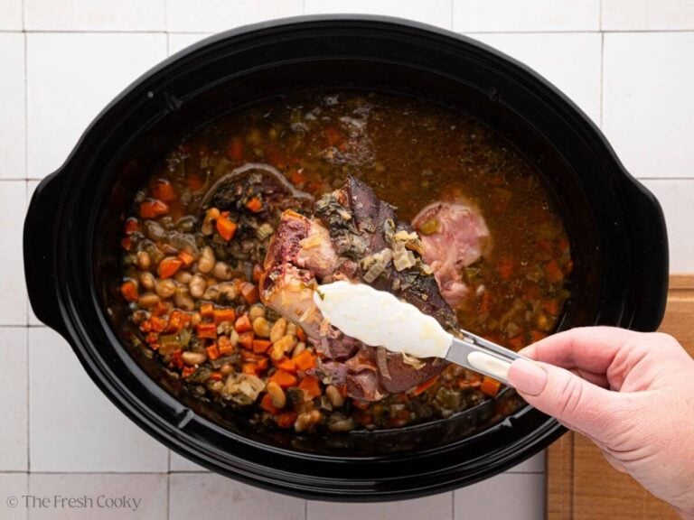 A black slow cooker bowl with ham and bean soup. Tongs are holding up a ham hock with a hand.