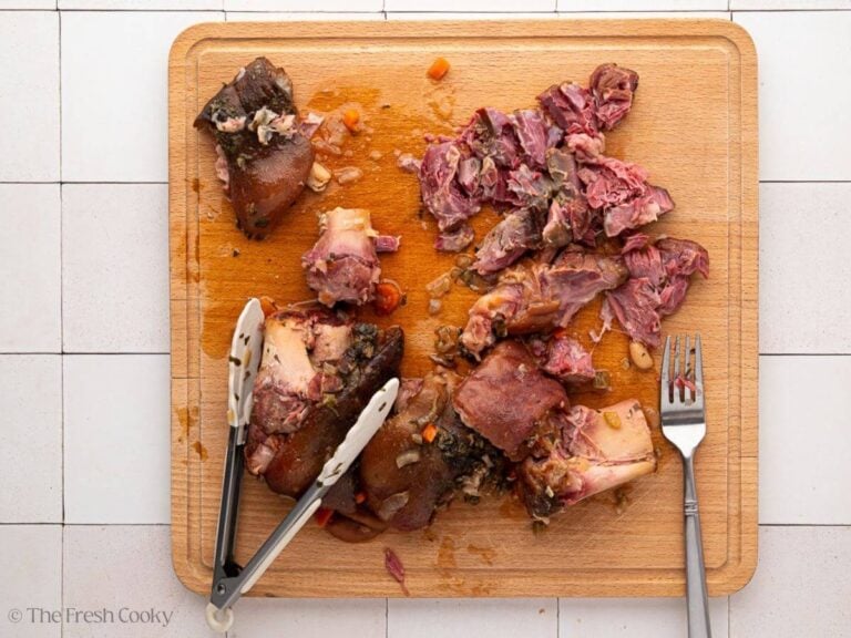 A wooden cutting board with shredded ham from a ham hock with a fork and tongs.