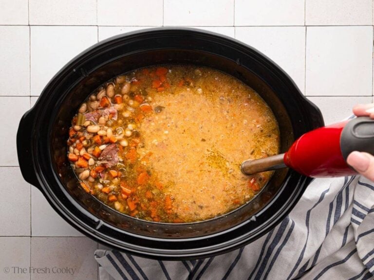 A black slow cooker bowl with slow cooker ham and bean soup with an immersion blender and a hand.