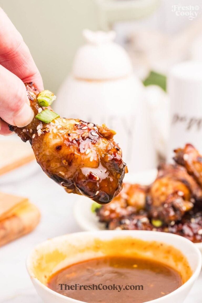 Holding a drummette sticky chicken wing with other wings in background and a dipping sauce below, to Pin.