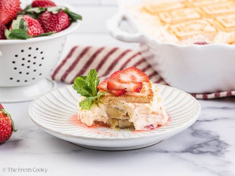 A white plate of strawberry banana pudding with a baking dish and bowl of strawberries in the background.
