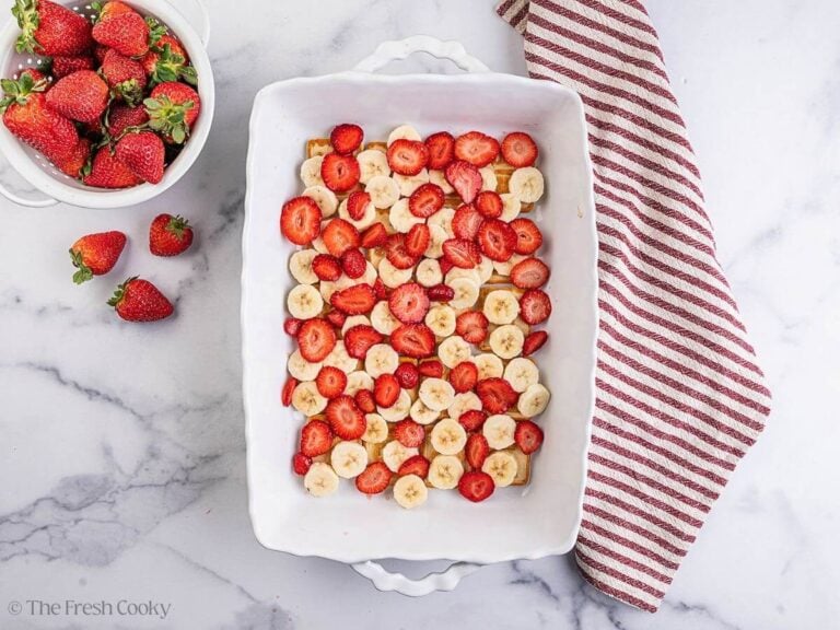 A white baking dish with cookies, sliced bananas and strawberries in the bottom. A white bowl of strawberries in to the side and a striped linen.