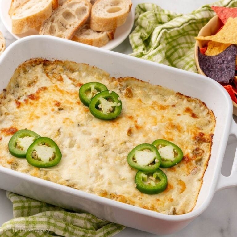 Baked artichoke jalapeno dip in a white baking dish next to bread and chips.