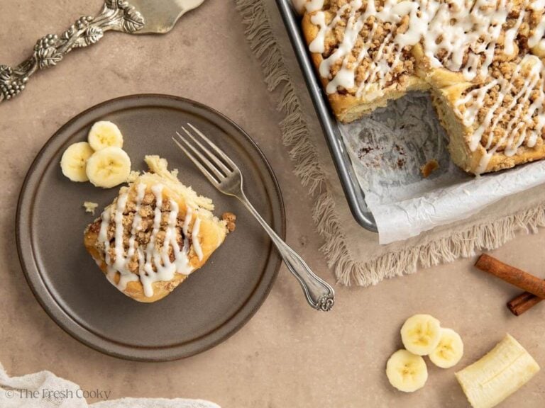 Banana bread cinnamon rolls in pan with one roll removed to a gray plate with a fork and sliced bananas.