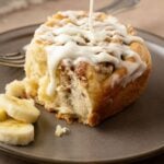 Banana Bread cinnamon roll on a gray plate, drizzling with glaze and sliced bananas on the side.