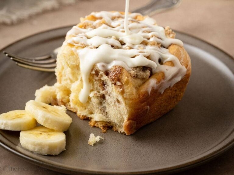 Banana Bread cinnamon roll on a gray plate, drizzling with glaze and sliced bananas on the side.