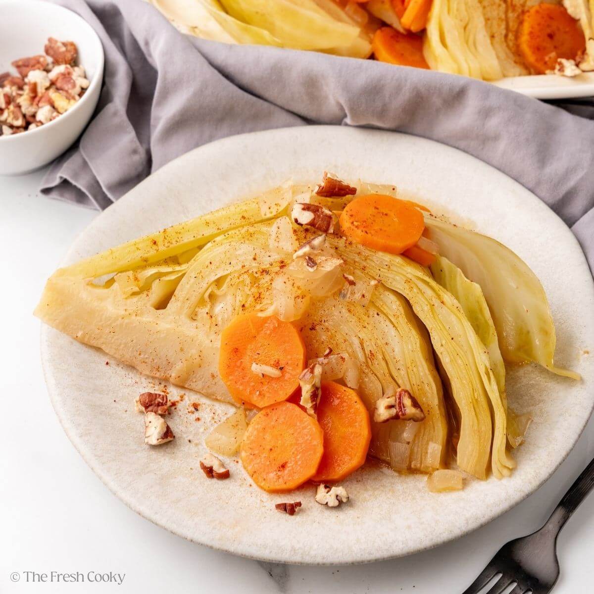 Braised cabbage wedge served on a plate with carrots and pecans on top.
