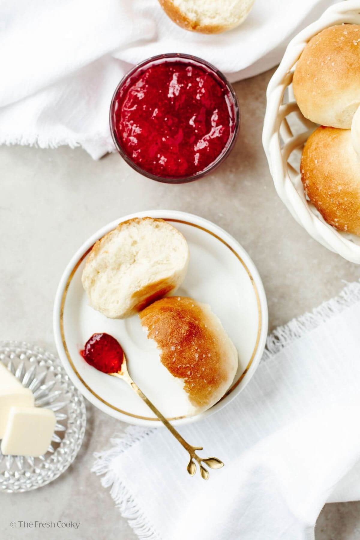 Yeast roll broken open on a plate with a basket of rolls nearby and a bowl of jam. 