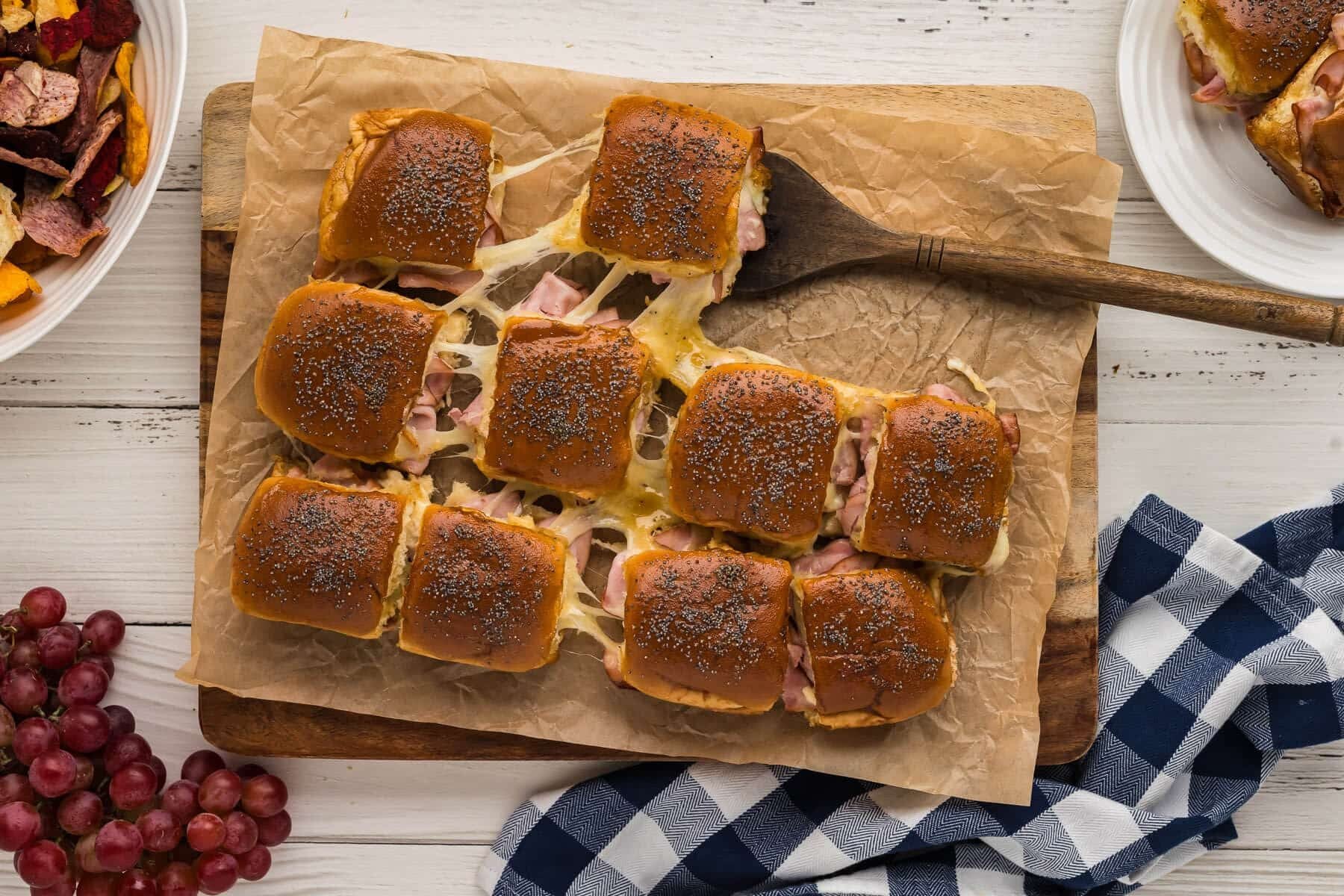 A bunch of ham and cheese sliders on a cutting board lined with parchment paper and pulling one out with a serving spatula.