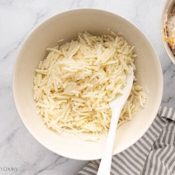 A white bowl with shredded frozen hash browns with a white rubber spatula. A second bowl is popping in on the right side with the ham, onion, sour cream, and cheese mixture with a wooden spoon.