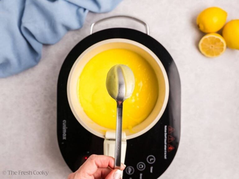 A hand holding a spoon with thickened lemon curd over a saucepan of finished curd.