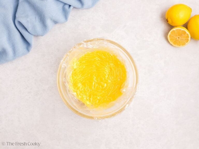 A small glass bowl with lemon curd with plastic wrap over top.