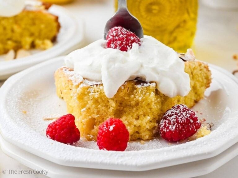 A white plate with a slice of almond flour olive oil cake with a fork about to take a bite.