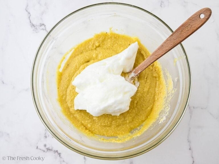 A glass bowl with almond flour olive oil cake batter with beaten egg whites on top. A wooden spoon is resting in the bowl.