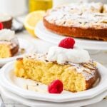 A slice of almond flour olive oil cake on a white plate with the whole cake in the background.