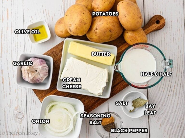 Labeled ingredients for garlic mashed potatoes L-R whole garlic bulb, olive oil, yukon gold potatoes, half and half, salt, bay leaf, seasoning salt, black pepper, sliced onion, cream cheese and butter.