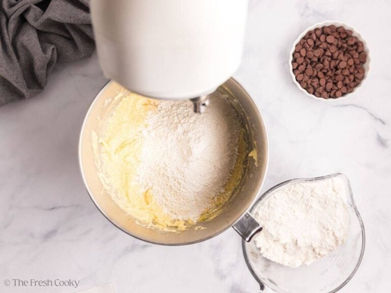Overhead image of a stand mixer with flour being added to vanilla cake batter. A glass bowl with excess flour is to the right.