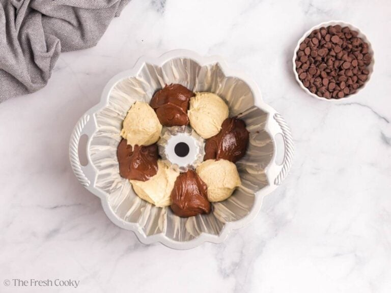 A bundt pan with scoops of vanilla and chocolate cake batter. A white bowl of chocolate chips is to the side.