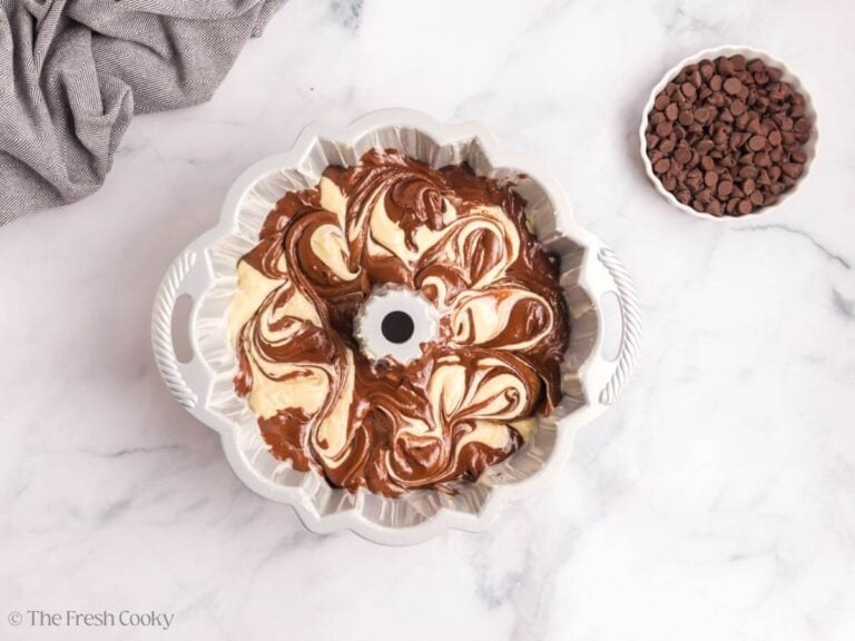A bundt cake pan with marble cake batter and a white bowl of chocolate chips to the side.