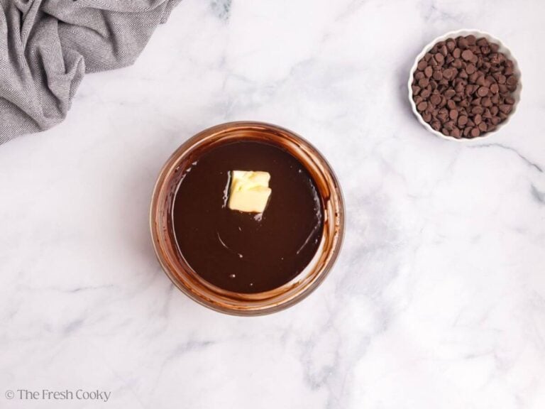 A glass bowl with chocolate ganache with two pats of butter on top. A white bowl with chocolate chips is to the right.