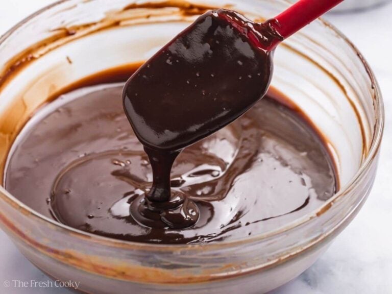 A red spatula is lifting up chocolate glaze in a glass bowl.