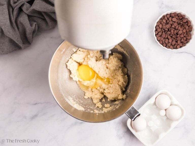 Overhead image of a stand mixer with wet ingredient creamed and eggs being added. A white container of eggs is to the side.