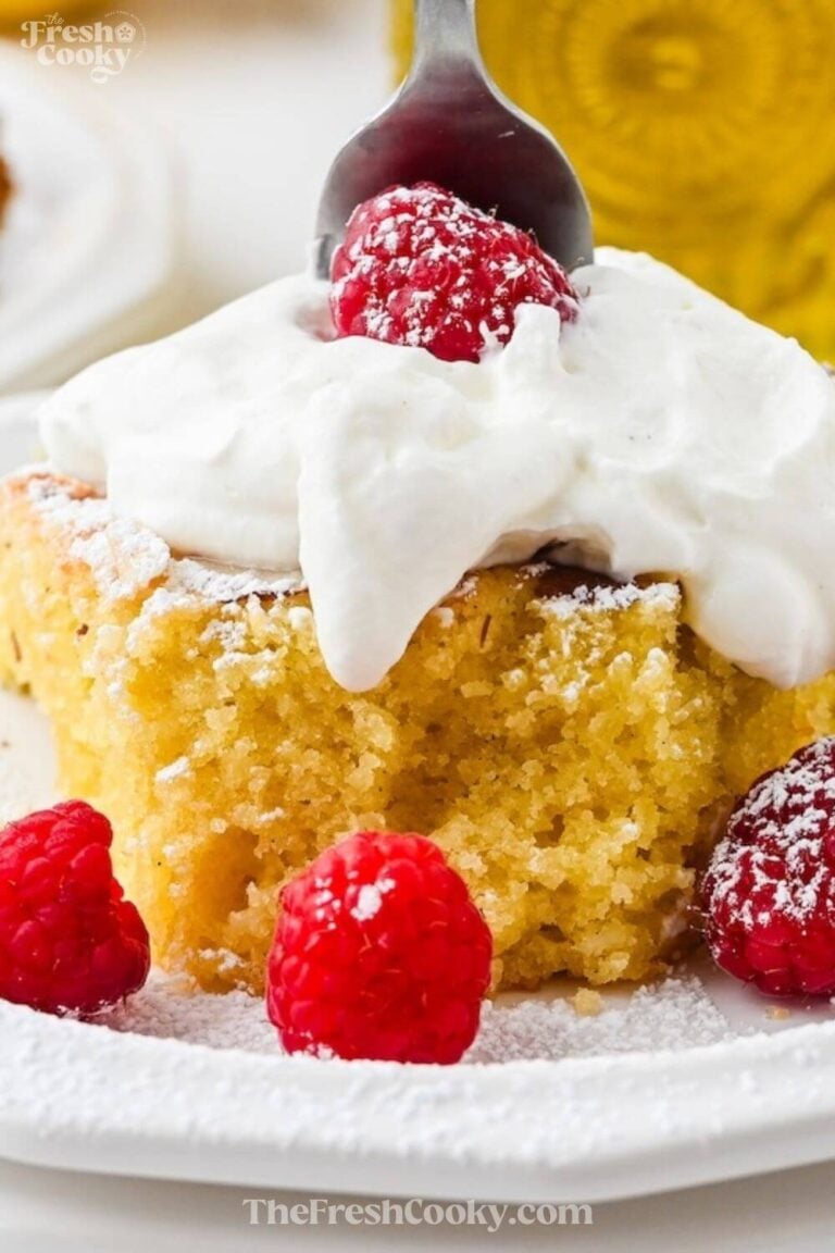 A slice of olive oil cake topped with whipped cream and surrounded by raspberries, to pin.