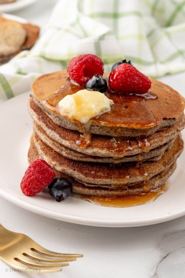 A white plate with a stack of buckwheat pancakes with syrup, butter, and berries on top.