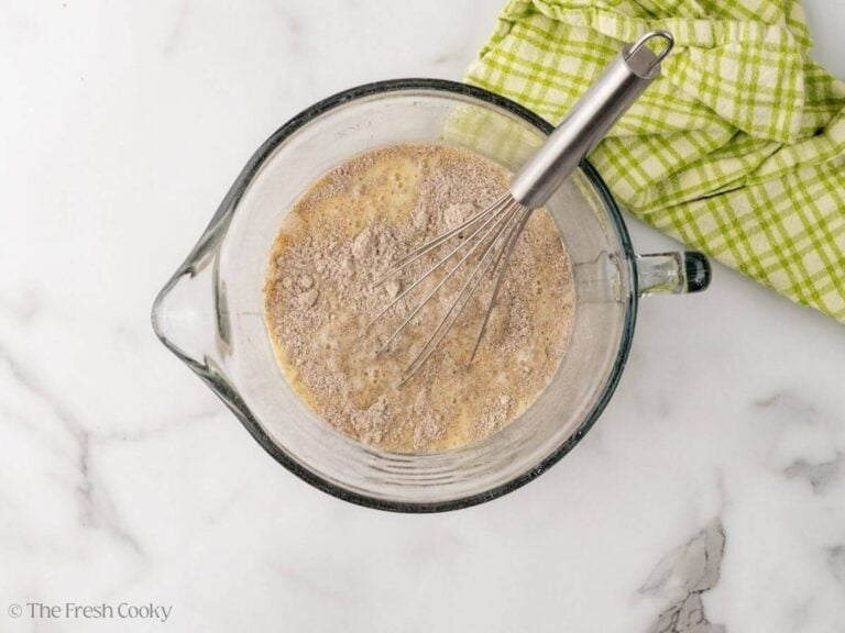 A large glass measuring cup with dry ingredients whisking into the wet ingredients. A whisk is resting in the large measuring cup.