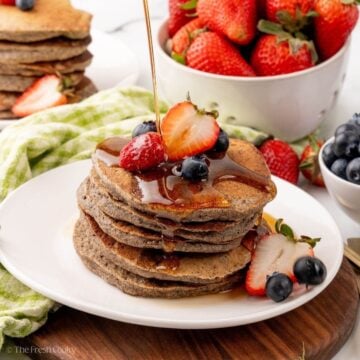 A white plate with a stack of buckwheat pancakes. Berries are on top with syrup drizzling over.