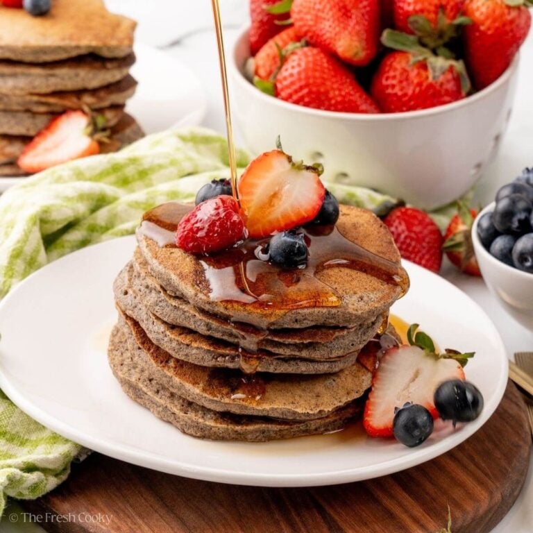 A white plate with a stack of buckwheat pancakes. Berries are on top with syrup drizzling over.