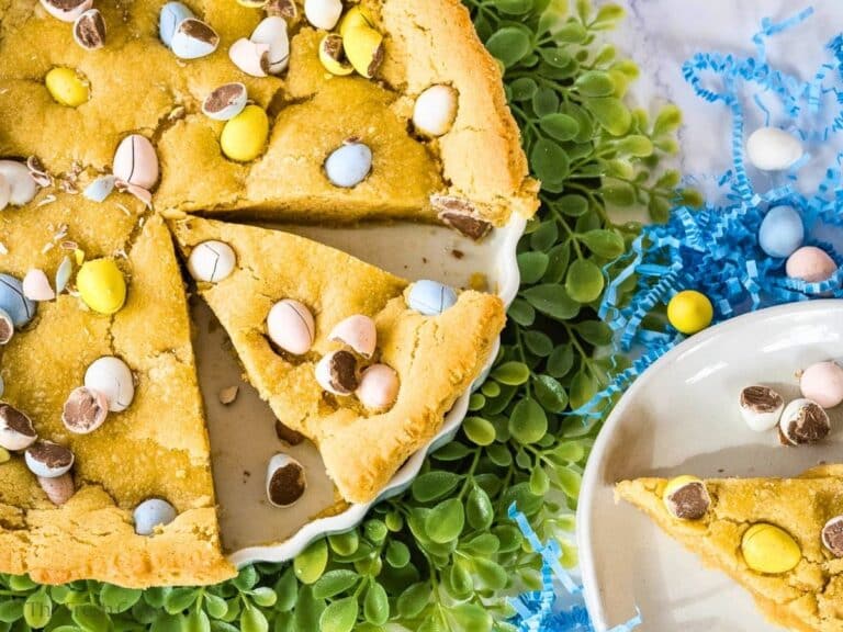 A giant Cadbury egg sugar cookie cake dotted with chocolate Cadbury Egg candies, with a slice removed and in the pan.