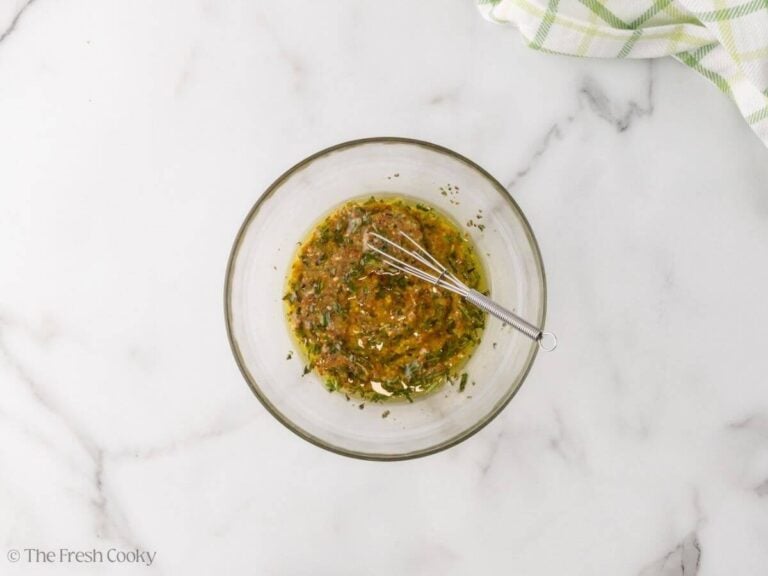 A glass bowl with the mustard, olive oil and herbs whisked together with a whisk resting on the side.