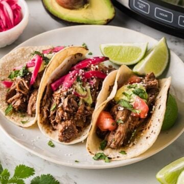 Barbacoa tacos with toppings on a plate surrounded by avocado, limes and pickled red onions.