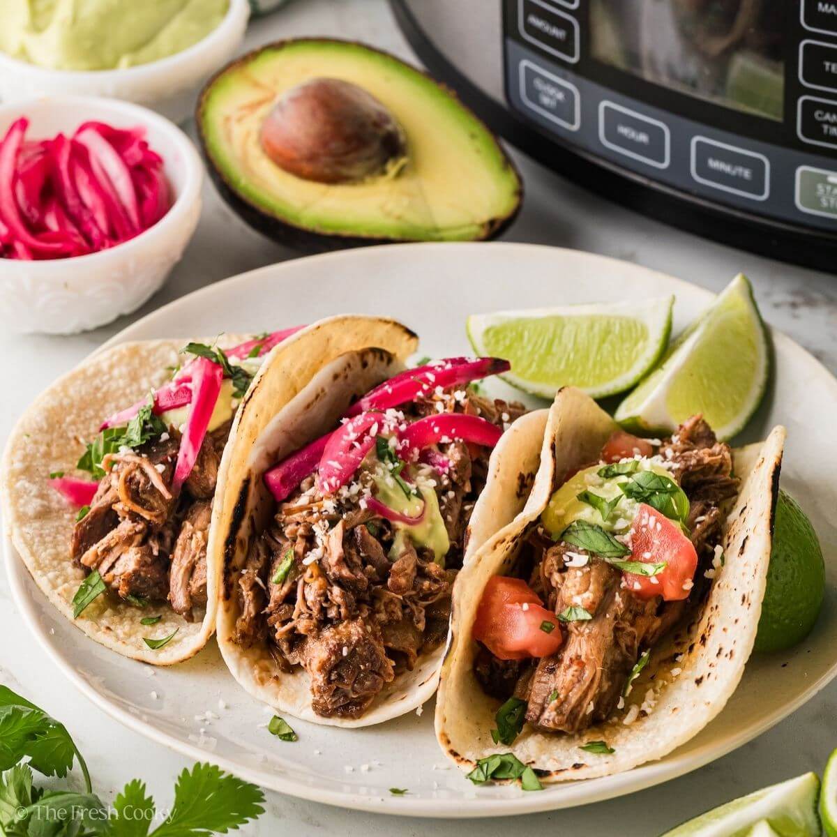 Slow cooker behind, with three barbacoa beef tacos on a plate, with limes, pickled red onions and crema.