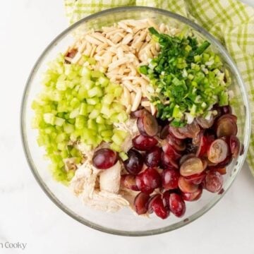 Celery, red grapes, shredded chicken, slivered almonds and green onions in glass bowl.