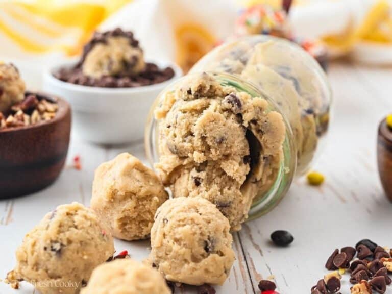 A spilled glass jar filled with edible chocolate chip cookie dough balls, with mix-ins in little bowls around it.