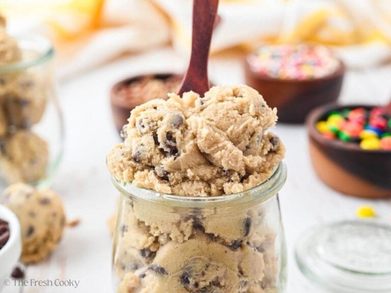 A jar of edible cookie dough with a wooden spoon in it and toppings behind.