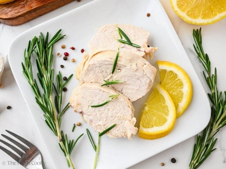 Three slices of poached chicken on a plate with rosemary and lemon slices on the side.